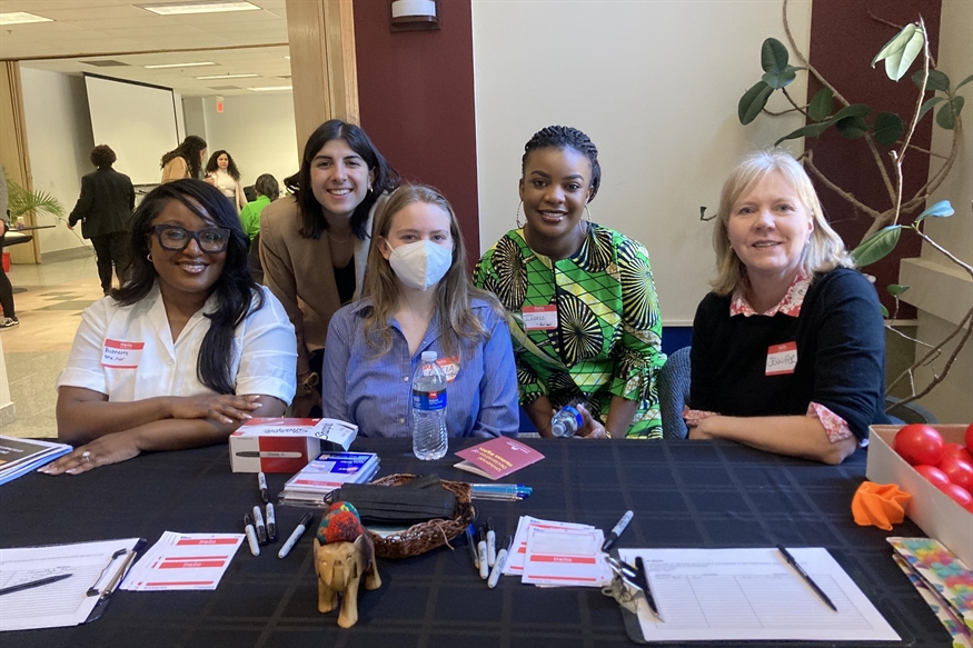 [ai] Five women seated at a registration table during an event, with various papers and a water bottle visible. The setting features a modern interior, and participants in the background are engaged in conversation.